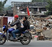 Pora važiuoja motociklu &scaron;alia sugriuvusių pastatų.