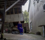 Moteris sėdi &scaron;alia namo, kuris buvo apgadintas per žemės nuo&scaron;liaužą, o &scaron;alia stovinitis naujas namas pradėjo smegti.