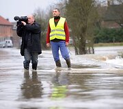 Žurnalistai stovi ant apsemto kelio