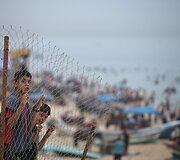 Palestiniečių berniukai už tvoros stebi žmones besimėgaujančius Viduržemio jūros paplūdimiu