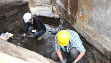 Klaipėdos universiteto archeologai, tyrinėjantys piliavietės teritoriją, aptiko XIII-ame amžiuje statyto medinio namo sienojus. Kairėje matomi gerai i&scaron;silaikę mediniai rastai. 