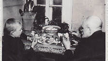Fotonuotrauka. Senelis (de&scaron;inėje) su anūku (kairėje) dažo velykinius margučius prie uždaroje patalpoje esančio stalo. XX a. 3&ndash;4 de&scaron;. (Trakų muziejus, CC BY)