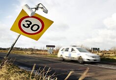 Policijos automobilis važiuoja pro vėjo nulenktą kelio ženklą.