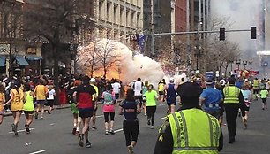 Bostono maratono dalyviai bėga link finišo, kairėje pusėje matomas prasidėjęs sprogimas.