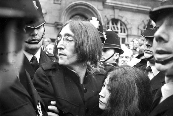 „Scanpix“ nuotr./Johnas Lennonas saugo Yoko Ono nuo minios prie Londono teismo, kuriame buvo apkaltinti marihuanos laikymu ir pasipriešinimu policijai.
