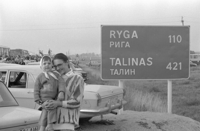 Lietuvos centrinio valstybės archyvo (toliau – LCVA) fondų nuotrauka./Fotografas, žurnalistas iš Klaipėdos Antanas Stanevičius fotografuoja Baltijos kelio akcijos žmones ruože prie Pasvalio. Kelio ženklas, informuojantis apie atstumus iki Rygos ir Talino. Stovi lituanistė Eglė Zalatoriūtė su dukra Elvyra. 