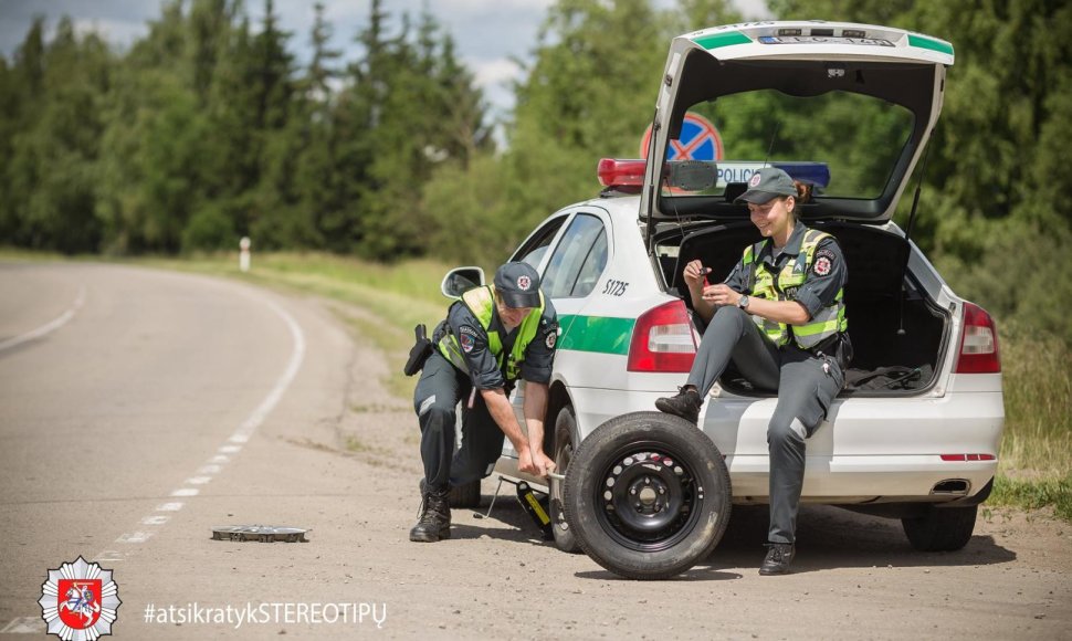 Akcijos „Atsikratyk stereotipų“ fotosesijos kadras: kol kolega keičia tarnybinio automobilio ratą, jo porininkė lakuojasi nagus