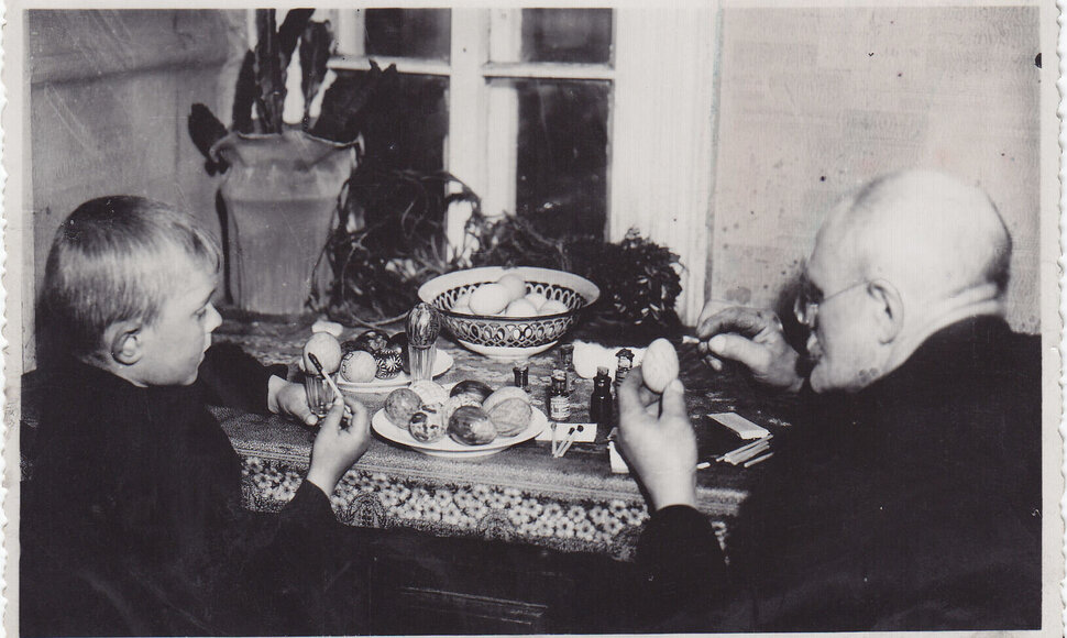 Fotonuotrauka. Senelis (de&scaron;inėje) su anūku (kairėje) dažo velykinius margučius prie uždaroje patalpoje esančio stalo. XX a. 3&ndash;4 de&scaron;. (Trakų muziejus, CC BY)